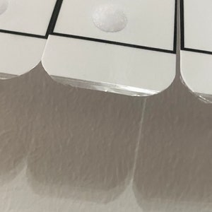 以下が含まれることがあります： 丸みを帯びた角と黒い縁取りのある、白い長方形のカードのクローズアップ。各カードには、小さく丸い白い粘着ドットが付いています。カードは部分的に透明なプラスチックで覆われています。