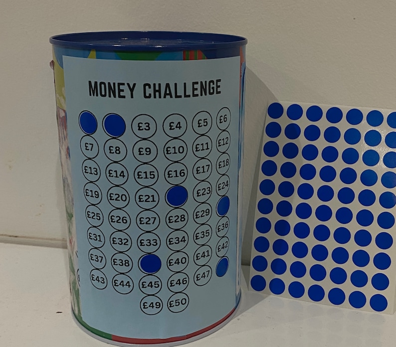 May include: A blue and white money challenge jar with a grid of numbers from 3 to 50. The jar has a blue lid and a blue sticker with the words "MONEY CHALLENGE" on it. The jar is filled with blue stickers that have numbers on them.