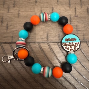 May include: A beaded keychain wristlet with a mix of orange, black, turquoise, and striped beads. A charm with the word "mama" in script is attached. The wristlet has a silver clasp.