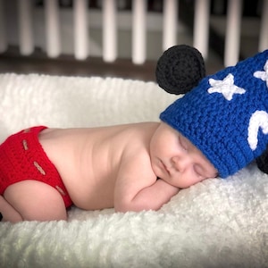 Peut inclure: Un bébé endormi portant un bonnet bleu crocheté avec des étoiles blanches et une lune croissante, et des oreilles noires. Le bébé porte une couche rouge en crochet avec des bordures noires.