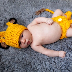 May include: A baby wearing a yellow crocheted lion costume with a hat, trousers, and booties. The costume has brown ears and a tail.