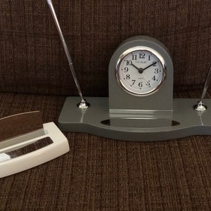 May include: Gray desk organizer with a clock, two pen holders, and a business card holder. The clock face is white with black numbers and the text "DANBURY" in black.