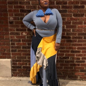 Peut inclure: Une personne portant un haut bleu à manches longues avec un style enroulé et une jupe en patchwork jaune, bleu et gris. La personne porte un turban à motifs et de grandes boucles d'oreilles à glands bleus.
