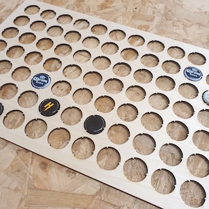 May include: A light-coloured wooden display board with numerous circular cutouts designed to hold bottle caps. Several bottle caps are visible, including a black cap and a Corona Extra cap. The board is ideal for showcasing a bottle cap collection.
