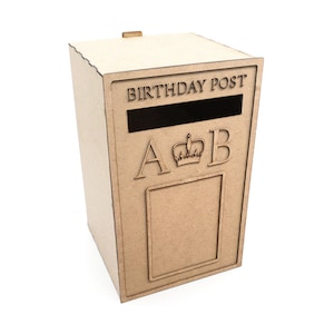 May include: A light brown wooden birthday post box with the words "BIRTHDAY POST" above a slot. The letters "A" and "B" flank a crown, and a rectangular frame is below. The box is on a white background.