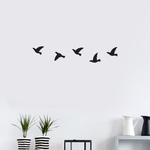 May include: A wall decoration featuring five black bird silhouettes in flight. The birds are arranged in a horizontal line against a white background. The image includes two potted plants with black and white striped pots.