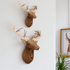 May include: Two decorative deer head wall hangings. The deer heads are made of layered, light brown wood, with detailed antlers. The heads are mounted on a white wall, creating a rustic and natural aesthetic. The design is modern and suitable for home decor.