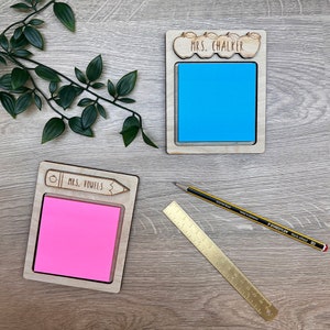 May include: Two wooden sticky note holders with teacher-themed designs. One has apples and the text "MRS. CHALKER", the other has a pencil and the text "MRS. VOWELS". A pencil and a brass ruler are also present.