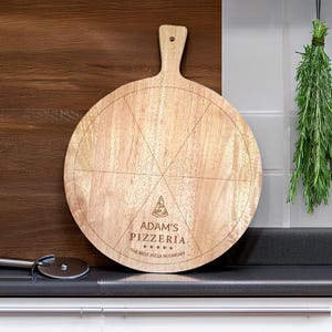 May include: A round wooden pizza serving board with a handle. The board is light brown and features etched lines for pizza slices and the text "Adam's Pizzeria" with a pizza slice graphic. The text "The Best Pizza in Cardiff" is also included.