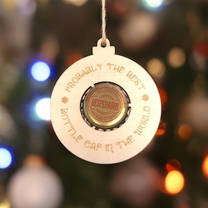 May include: A circular light-coloured wooden ornament featuring a central Desperados beer bottle cap. The wood is engraved with the text "PROBABLY THE BEST BOTTLE CAP IN THE WORLD" and includes a small twine loop at the top for hanging purposes.