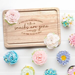 May include: A wooden cutting board with the text "IF YOU CAN READ THIS DANNY the snacks are gone IT IS TIME TO GO TO BED!" is surrounded by several flower-shaped cupcakes in various colors, including pink, blue, and yellow.