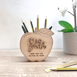 May include: Wooden apple-shaped desk tidy with the name "Mrs. Smith" and the message "Thank you for helping me grow" engraved on the front. The tidy holds pencils and pens. The apple is made of light-coloured wood.