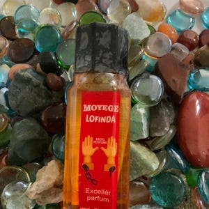 Pu&ograve; includere: Una bottiglia arancione trasparente con un tappo nero e un'etichetta rossa. L'etichetta dice "Moy&eacute;ge Lofinda Excel Perfume" e presenta un'immagine di due mani ammanettate. La bottiglia dice anche "Excell&eacute;r parfum" e "150ml".