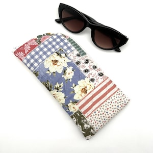 Puede incluir: Un estuche para gafas acolchado con estampado floral y cierre de cremallera. El estuche está hecho de un tejido suave y tiene un diseño de patchwork. El estuche se muestra con unas gafas de sol negras de ojo de gato.