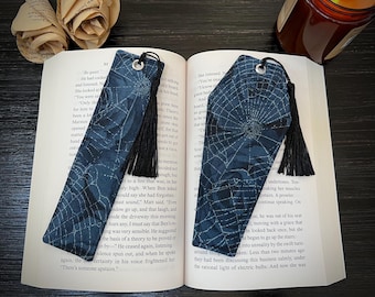 May include: Two fabric bookmarks with a dark blue spiderweb design and black tassels. One bookmark is rectangular, the other is coffin-shaped. Both are placed on an open book with text. A candle and decorative paper flowers are in the background.