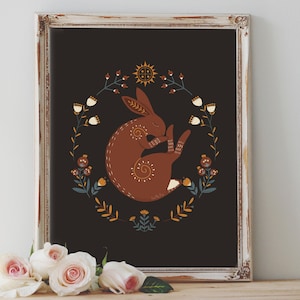 May include: Framed art print featuring a brown rabbit curled up in a floral wreath on a black background. The artwork includes a sun and floral elements in yellow, white, and blue. The frame is white with a distressed finish.