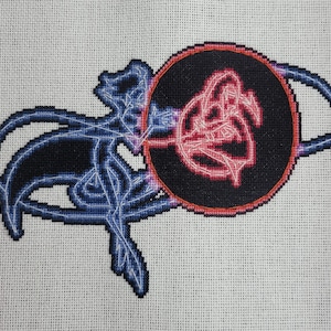 May include: Cross-stitch embroidery design featuring a stylised floral motif in pink and white, surrounded by a black circle with a white outline. The design is set against a white background.