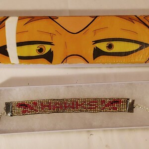 May include: A red and gold beaded bracelet with the words "PLUS ULTRA" in red beads. The bracelet is in a white box with a cartoon image of a character's eyes on the lid.