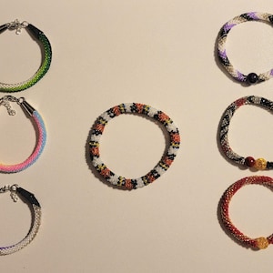 May include: A collection of beaded bracelets in various colors, including green, purple, red, and rainbow hues. Each bracelet is a closed loop, with a clasp closure. The bracelets are displayed on a light-colored surface.