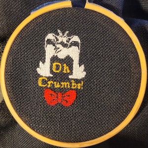 Peut inclure: Art de broderie sur un cercle représentant un personnage de dessin animé blanc avec le texte "Oh Crumbs!" et un nœud papillon rouge.
