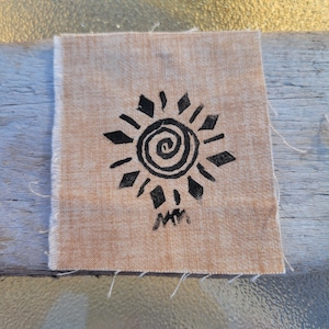 May include: A square piece of light tan fabric featuring a black ink print of a spiral sun design with diamond-shaped rays. Below the sun graphic, there is a small, hand-drawn black signature consisting of the letters MM in a stylized script.