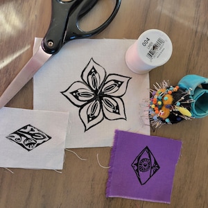 May include: Black scissors, a spool of light pink thread labeled 004 Linen, and a round pin cushion with colorful pins. Three fabric swatches are present: a large white square with a black flower print, a small white rectangle, and a purple square.