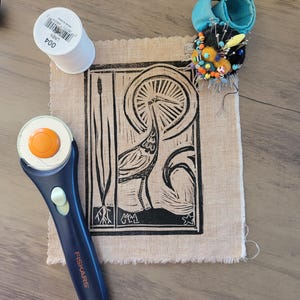 May include: A square piece of tan fabric featuring a black block print of a heron standing in water with a sunburst background. The fabric is accompanied by a white spool of linen thread, a blue rotary cutter, and a round pincushion with colorful pins.