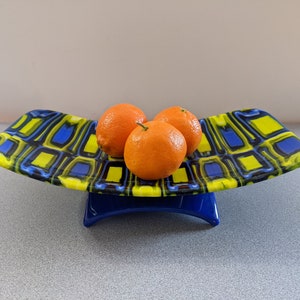 May include: A decorative glass fruit bowl with a blue base and a patterned top in yellow and blue. The bowl holds three oranges. The bowl has a unique, curved shape and is a stylish addition to any kitchen or dining room.