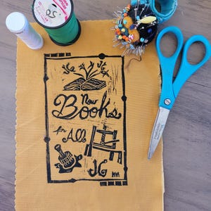 May include: A rectangular piece of mustard yellow fabric featuring a black block print design with an open book, a printing press, and the text "New Books for All JG". The fabric is surrounded by a spool of green thread, a white thread spool, a blue pincushion with colorful pins, and blue-handled Fiskars scissors.