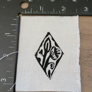 May include: A square piece of off-white fabric featuring a black diamond-shaped block print of a floral motif. A black metal L-shaped ruler is positioned along the top and left edges, showing the fabric measures approximately 2 inches wide.