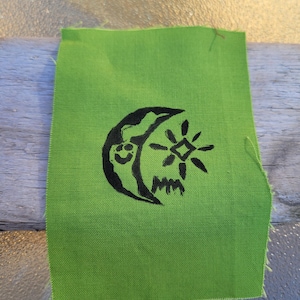 May include: A rectangular piece of lime green fabric featuring a black screen-printed graphic. The design depicts a crescent moon with a smiling face, a stylized starburst shape, and the letters MM printed in a bold, hand-drawn black ink style.
