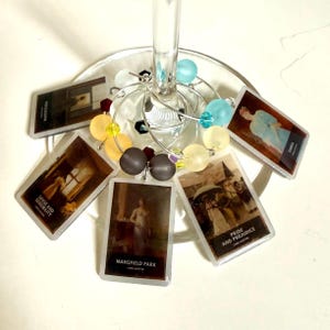 Op de afbeelding: Een set zilverkleurige wijnglasbedels met rechthoekige hangers met Jane Austen-boekomslagen zoals Mansfield Park, Pride and Prejudice en Sense and Sensibility, geaccentueerd met kleurrijke ronde kralen in blauw, geel en grijs.
