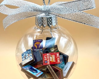 May include: A clear, spherical glass ornament filled with miniature book replicas. The ornament is topped with a silver metal cap and a shimmering silver ribbon tied in a bow. The tiny books inside feature various colorful covers and titles.