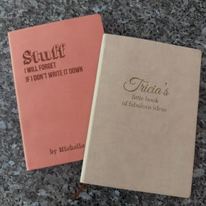 May include: Two notebooks are shown, one coral and one beige. The coral notebook has the text "Stuff I Will Forget If I Don't Write It Down" and "by Michelle." The beige notebook says "Tricia's little book of fabulous ideas."