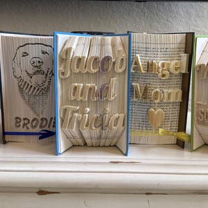 Könnte beinhalten: Fünf Buchfaltobjekte, jedes mit einem anderen Design. Das erste ist ein weißes Buch mit den Worten "Be the Light" in goldener Schrift. Das zweite ist ein weißes Buch mit einem Schwarzweißbild eines Hundes und dem Wort "Brodie" darunter. Das dritte ist ein weißes Buch mit den Worten "Jacob and Tricia" in goldener Schrift. Das vierte ist ein weißes Buch mit den Worten "Angel Mom" in goldener Schrift. Das fünfte ist ein weißes Buch mit den Worten "Welcome to So Booked Up" in goldener Schrift.