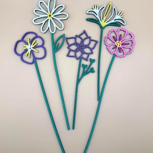 May include: A set of four flat, decorative flower stakes featuring cutout designs. The flowers include a white daisy, a purple pansy, a purple geometric blossom, and a pink cosmos, all attached to long, thin, teal green stems with leaf details.