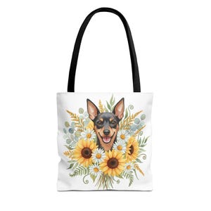 May include: White tote bag with a black handle featuring a watercolor illustration of a smiling black and brown dog surrounded by sunflowers and daisies. The dog has large ears and a happy expression. The bag is perfect for carrying groceries or personal items.