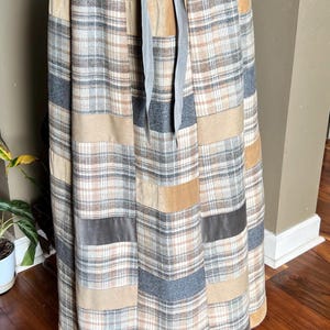 May include: A long skirt with a patchwork design. The skirt features a plaid pattern in shades of beige, gray, and brown, with velvet panels. It has a drawstring waist and a gray ribbon tie. The skirt is floor-length.