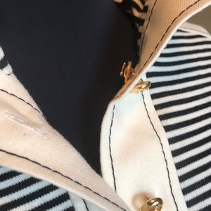 May include: Close-up of a white and black striped shirt with a gold button closure. The shirt has a collar and a black seam around the edges.