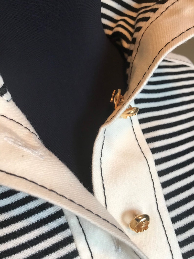 May include: Close-up of a white and black striped shirt with a gold button closure. The shirt has a collar and a black seam around the edges.