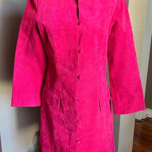 May include: A vibrant fuchsia-colored coat with a mandarin collar and long sleeves. The coat features a button-down front and two front pockets. The garment is displayed on a black mannequin, showcasing its tailored fit and design.