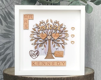 May include: A white framed picture with a wooden tree design inside. The tree has a heart with the words "I do" on it, and the date "281221" on the trunk. The names "Dale" and "Kayleigh" are on the branches, and the word "love" is on a heart at the bottom. The words "Mr & Mrs Kennedy" are spelled out with wooden tiles.