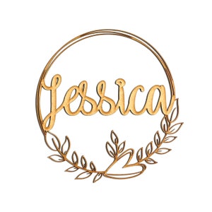 May include: A wooden sign with the name "Jessica" in cursive script, encircled by a gold-colored ring and adorned with a laurel wreath design. The sign is made of light-colored wood and is set against a white background.