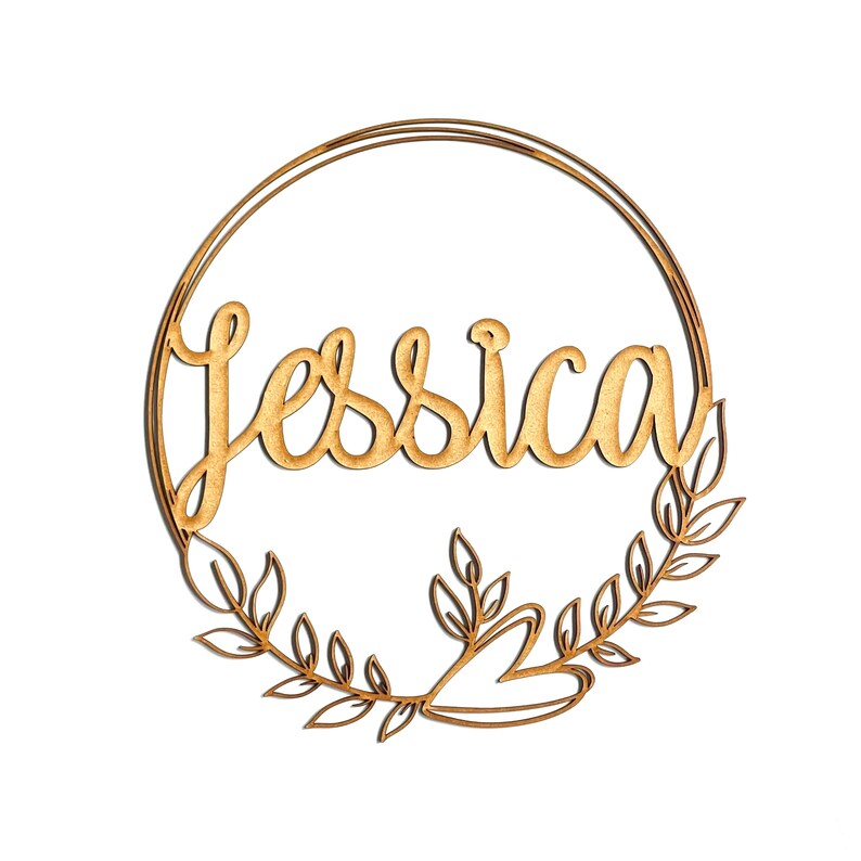 May include: A wooden sign with the name "Jessica" in cursive script, encircled by a gold-colored ring and adorned with a laurel wreath design. The sign is made of light-colored wood and is set against a white background.