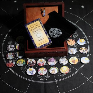 以下が含まれることがあります： 黒いベルベットの袋とタロットカードのデッキが入った木製の箱。箱が開いており、カードは白い円形のデザインが施された黒い布の上に広げられています。カードは丸く、カラフルな画像が描かれています。箱のテキストには「The Original Rider Waite Tarot Deck Compiled and Edited by A.E. Waite Cards Designed by Pamela Colman Smith.」と書いてあります。