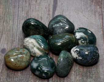May include: A collection of polished moss agate stones in shades of green, white, and brown. The stones have a smooth, rounded shape and vary in size. The background is a wooden surface. The website address www.WitchcraftEmporium.co.uk is visible.