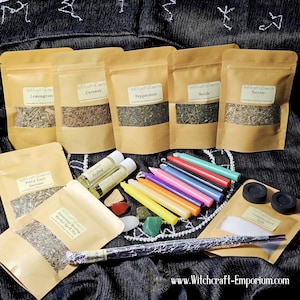 Op de afbeelding: Een verzameling gedroogde kruiden, wierook, kaarsen en andere magische benodigdheden voor hekserij en spreuken. De items zijn gerangschikt op een donkerblauwe stoffen achtergrond met de tekst "www.Witchcraft-Emporium.com".