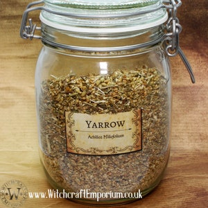 Puede incluir: Un frasco de vidrio lleno de hierba de milenrama seca, etiquetado como "Yarrow Achillea Millefolium". El frasco tiene una tapa plateada con un cierre.