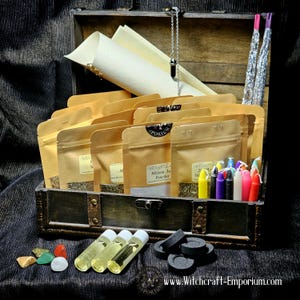 May include: A wooden treasure chest filled with various witchcraft supplies, including herbs, candles, oils, incense, and crystals. The chest is open and the items are arranged neatly inside. The text "www.Witchcraft-Emporium.com" is visible on the bottom right corner of the image.