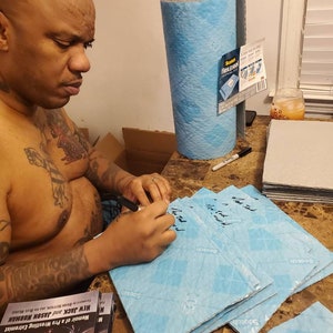 May include: A person signing a stack of blue Scotch Flex & Seal tape. The tape is labeled "Scotch Flex & Seal" and has a diamond pattern. The person is signing the tape with a black marker. The book "Memoir of a Pro Wrestling Extremist: New Jack and Jason Norman" is visible in the foreground.